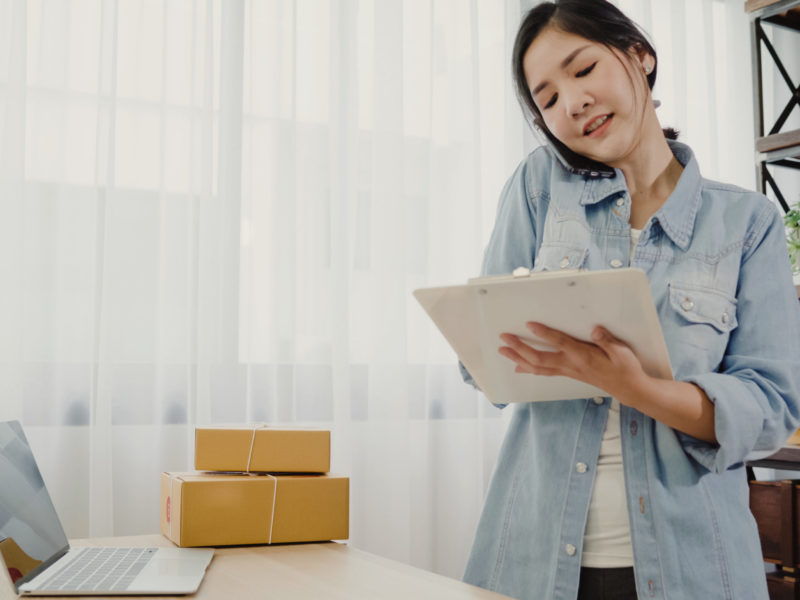 photo of woman owner of online small business talking on phone checking product stock and writing on clipboard