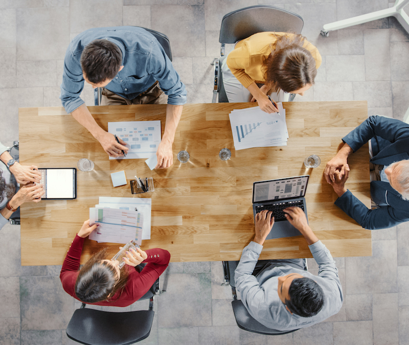 Startup Meeting Room: Team of Entrepreneurs sitting at the Conference Table Have Discussions, Solve Problems, Use Digital Tablet, Laptop, Share Documents with Statistics, Charts. Top View