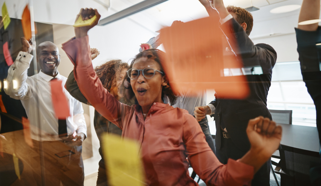 Diverse group of ecstatic businesspeople cheering together over a winning idea while brainstorming with sticky notes on a glass wall in a modern office