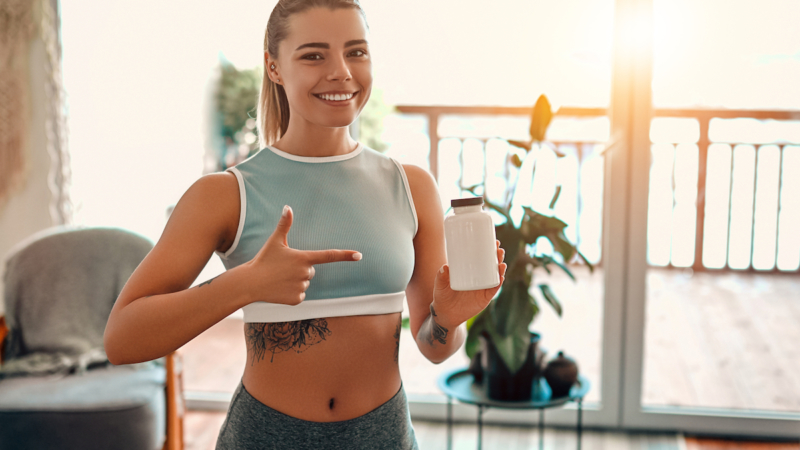 Athletic woman in sportswear holds a jar of protein in her hand and shows a finger gesture at home in the living room. Sport and recreation concept.