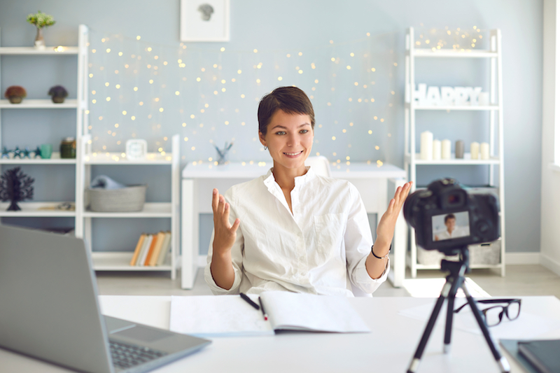 Positive female vlogger recording a video on camera sitting at desk in modern office. Happy businesswoman sharing experience making a pre-recorded webinar at home