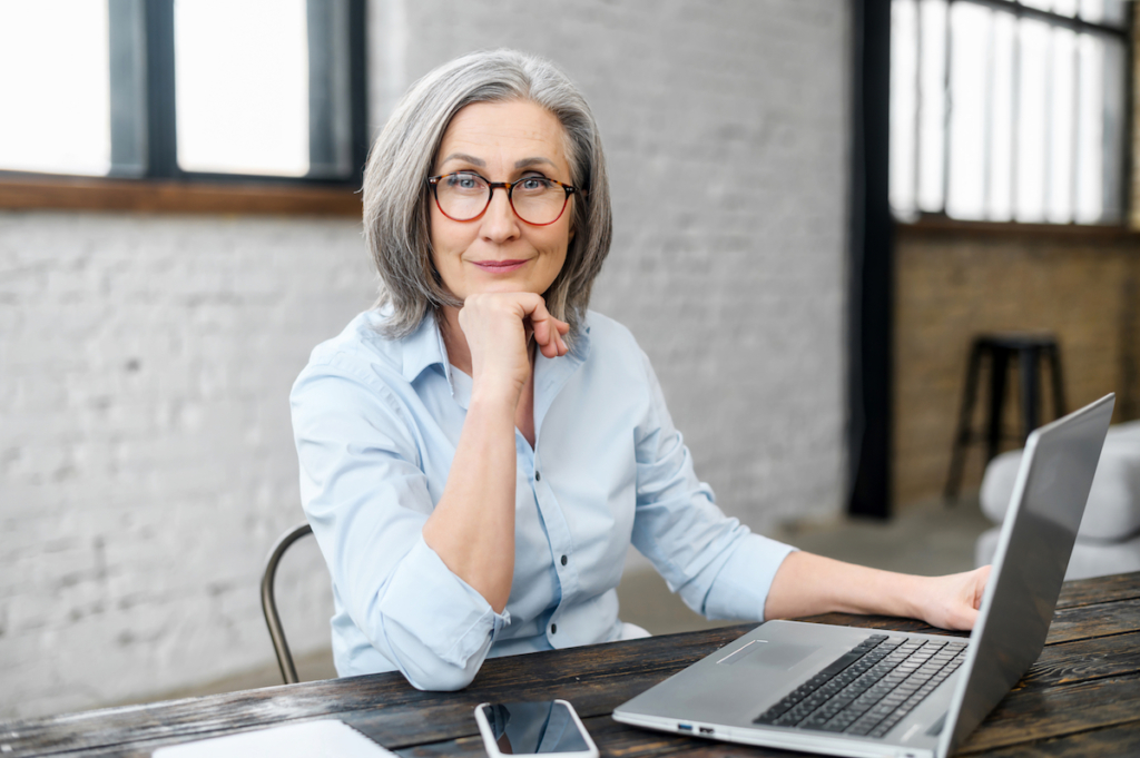 Intelligent mature senior female business owner in glasses with hand on a chin looking at the camera, sitting at the desk in front of laptop. 