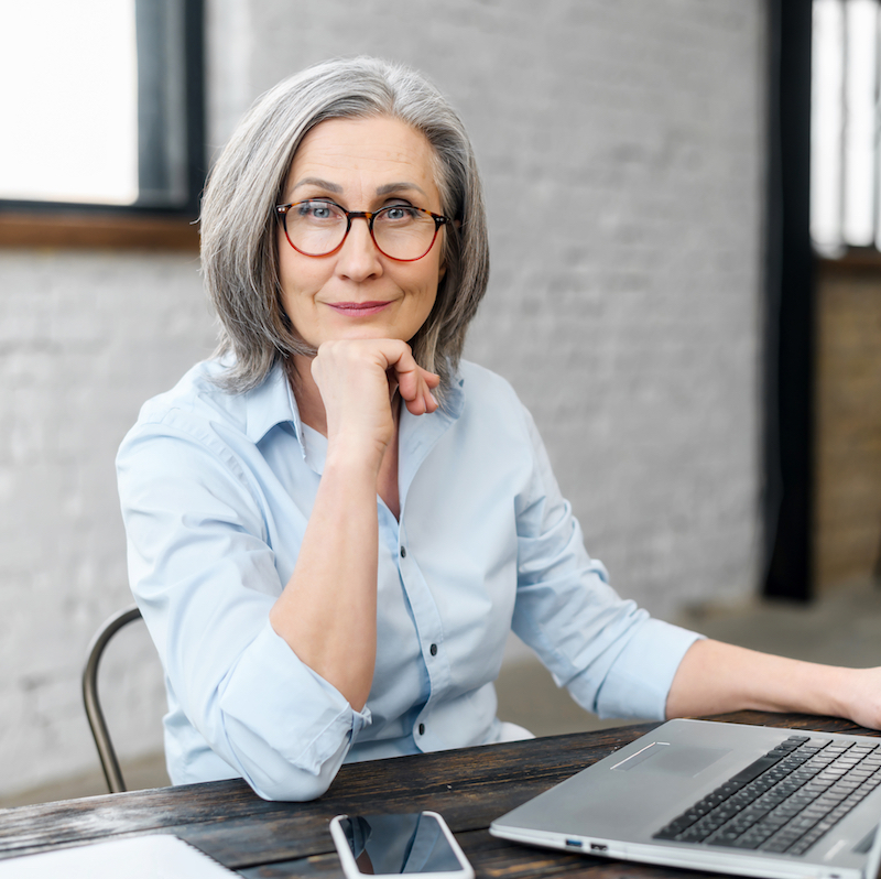 Intelligent mature senior female business owner in glasses with hand on a chin looking at the camera, sitting at the desk in front of laptop.