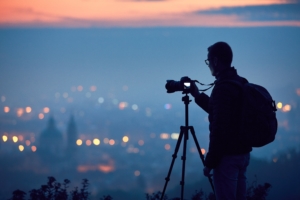 Silhouette of photographer with tripod on hilltop taking photos of city lights at night