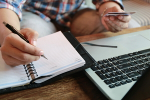 photo of man writing in a notebook and holding a credit card and laptop on table