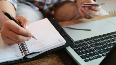 photo of man writing in a notebook and holding a credit card and laptop on table