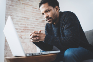 Young man thinking with hands folding looking at laptop screen