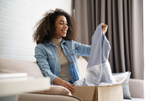 Young curly hair satisfied happy african girl woman lady shopaholic customer sit on sofa unpack parcel delivery box look at clothes take out grey pullover sweater, online shopping shipment concept.