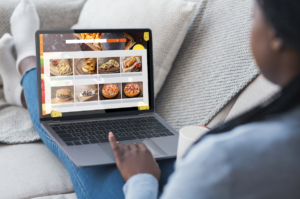 Black Woman Using Laptop Ordering Fast food Meals Online On Restaurant Website Sitting On Sofa At Home. 