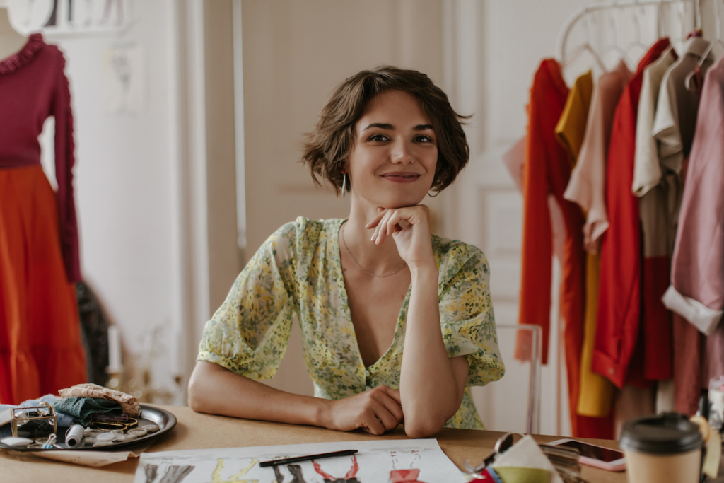 Attractive young curly short-haired woman in floral dress smiles and looks into camera. Portrait of fashion designer in office.
