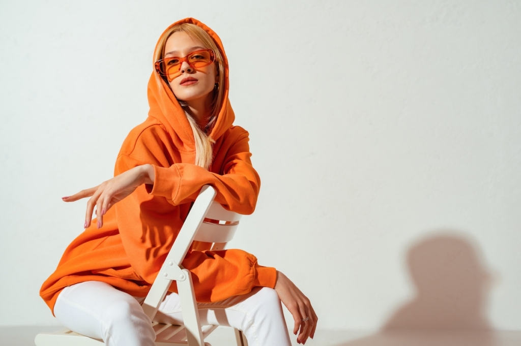 Fashionable confident blonde woman wearing trendy orange sweatshirt, color sunglasses, posing on white background. 