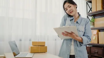 photo of woman owner of online small business talking on phone checking product stock and writing on clipboard