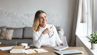 photo of woman author sitting at desk reading with books & laptop