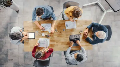 Startup Meeting Room: Team of Entrepreneurs sitting at the Conference Table Have Discussions, Solve Problems, Use Digital Tablet, Laptop, Share Documents with Statistics, Charts. Top View