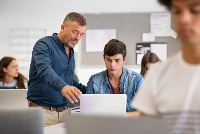 Professor Assisting College Student With Laptop In Classroom