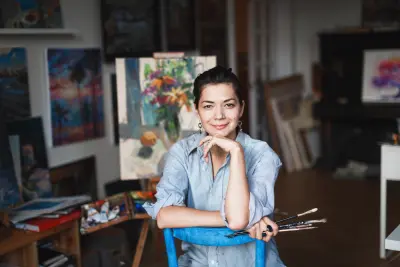 A young smiling brunette woman artist in her Studio is holding a brush. Near her easel, paintings and various art equipment.