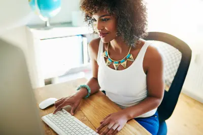 Stylish young African American entrepreneur working from her home office on her website sitting at a desk reading the screen of a large desktop monitor