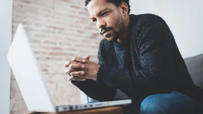 Young man thinking with hands folding looking at laptop screen