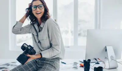 Smiling female photographer with a professional camera sitting on her desk. Woman with dslr camera in office looking away and smiling.