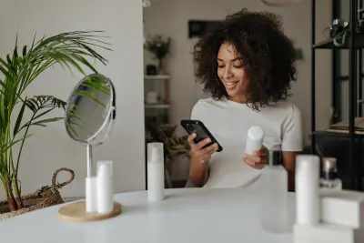 Cheerful brunette woman in white t-shirt smiles, reads recommendations in smartphone and holds great face cream bottle
