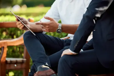 two people in business clothes sitting on a bench outside looking at tablet