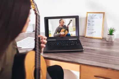 Focused girl playing acoustic guitar and watching online course on laptop while practicing at home. Online training, online classes.