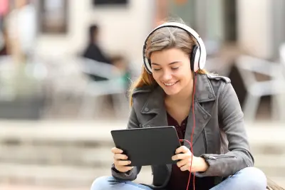 Beautiful fashion girl learning on line with a tablet and headphones sitting on a bench in the street